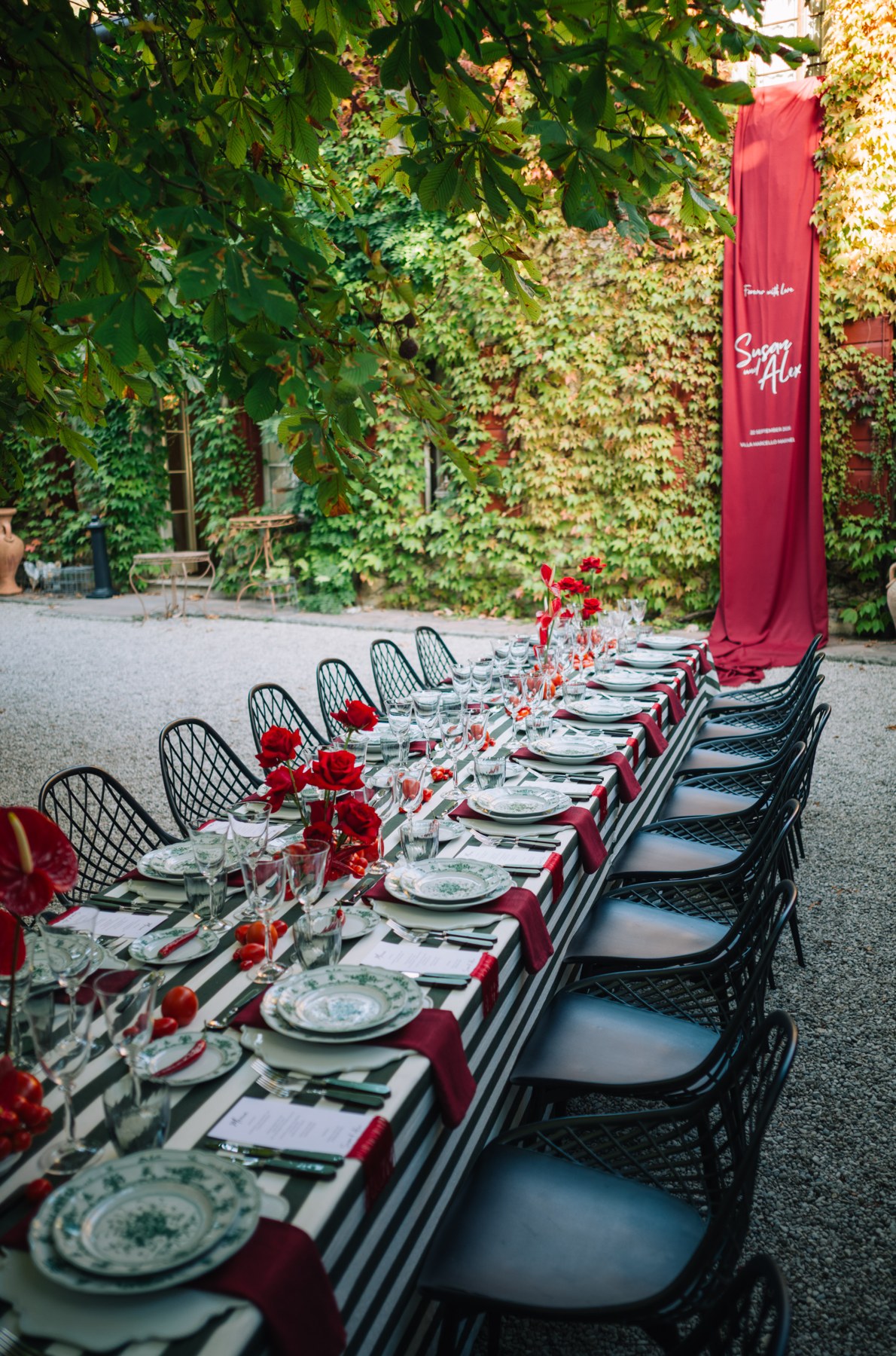 Lange gedeckte Tafel mit schwarzen Stühlen, gemustertem Geschirr und roten Blumen im Garten.