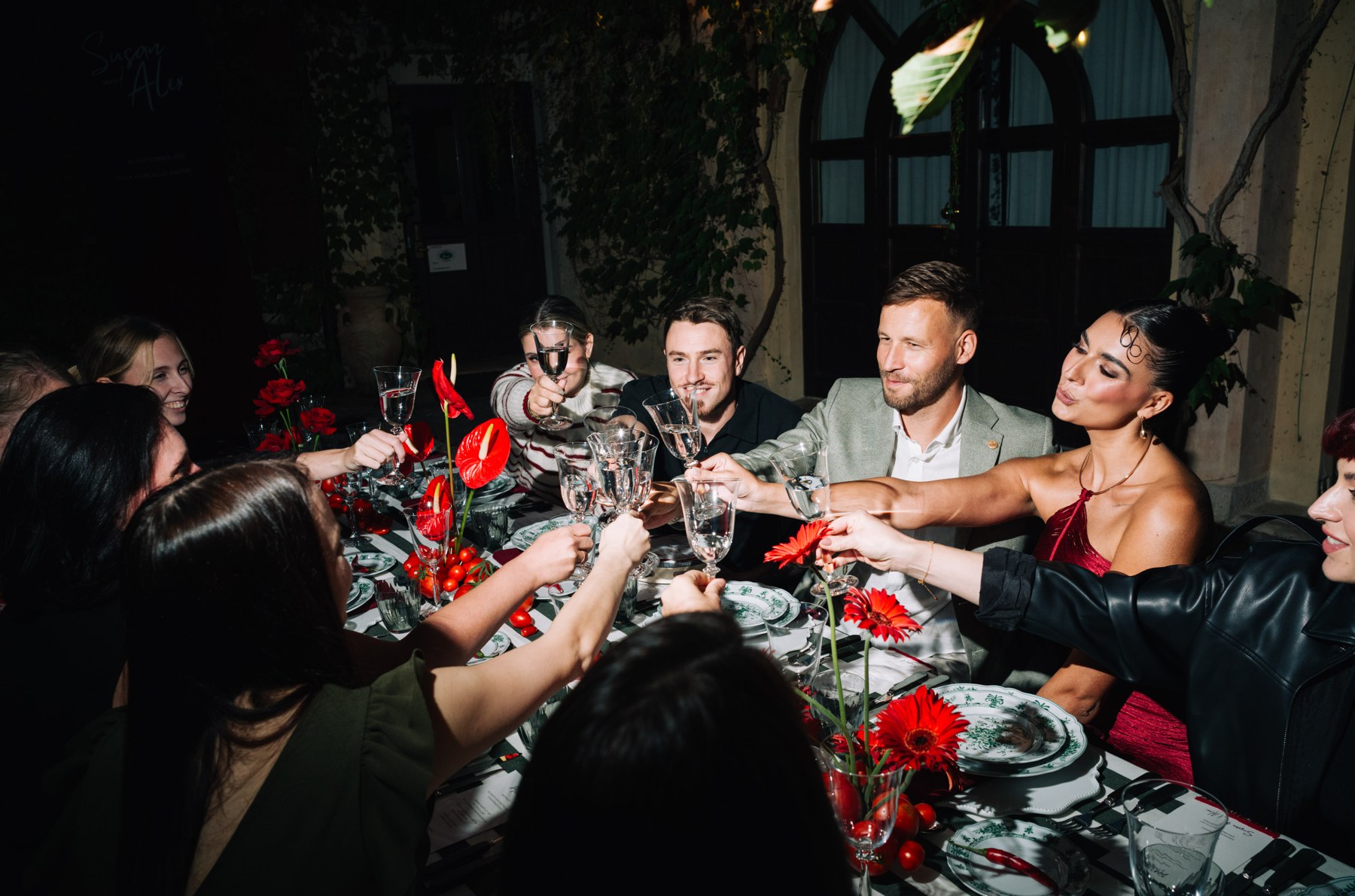 Mehrere Personen stoßen über einer mit roten Blumen dekorierten Dinner-Tafel an.