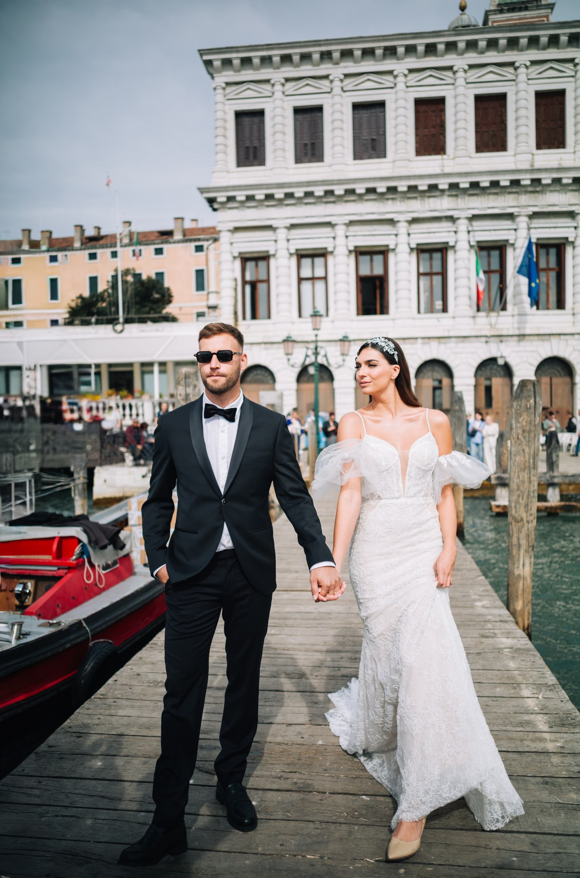 Paar in weißem Kleid und schwarzem Smoking geht am Wasser in Venedig entlang.