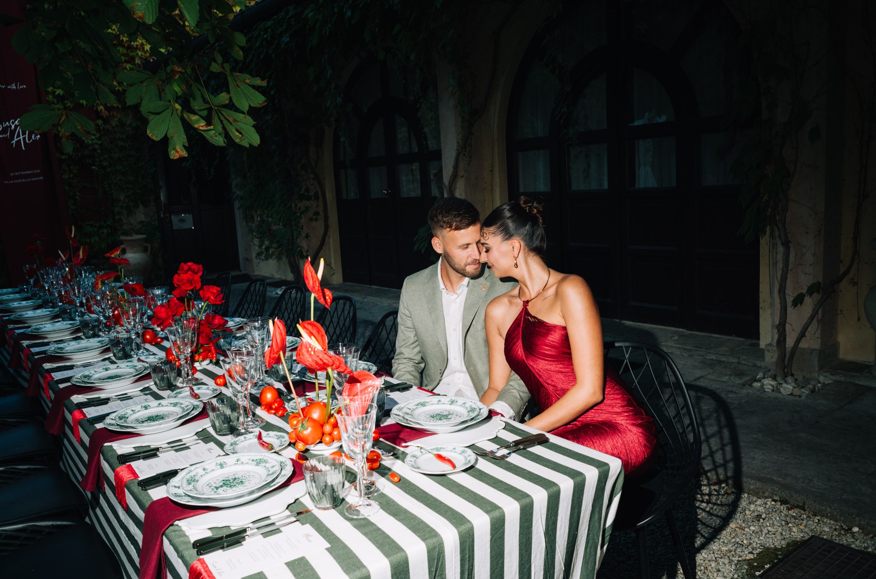 Paar sitzt eng beieinander an einer mit roten Blumen und gestreifter Tischwäsche gedeckten Tafel.